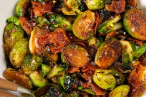 A white bowl filled with hot honey Brussels sprouts mixed with crispy bacon pieces and topped with chili flakes and flaky sea salt.