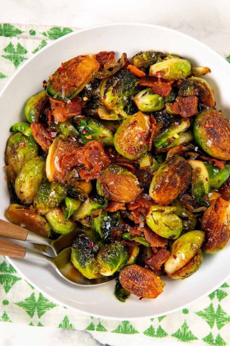A white bowl filled with hot honey Brussels sprouts mixed with crispy bacon pieces and topped with chili flakes and flaky sea salt.