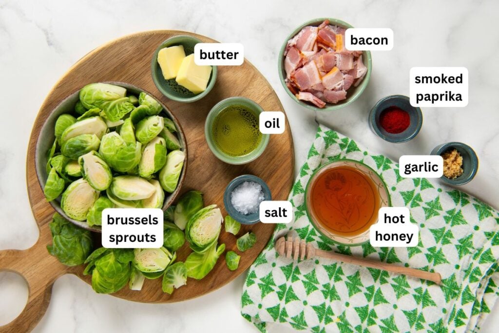 Hot Honey Brussels Sprouts with crispy bacon, caramelized sprouts, and a sweet-spicy honey drizzle. One skillet, ready in 25 minutes! Labeled ingredients for hot honey Brussels sprouts recipe arranged on a countertop: small bowls of butter, oil, salt, chopped bacon, smoked paprika, minced garlic, hot honey, and fresh Brussels sprouts sliced in half.