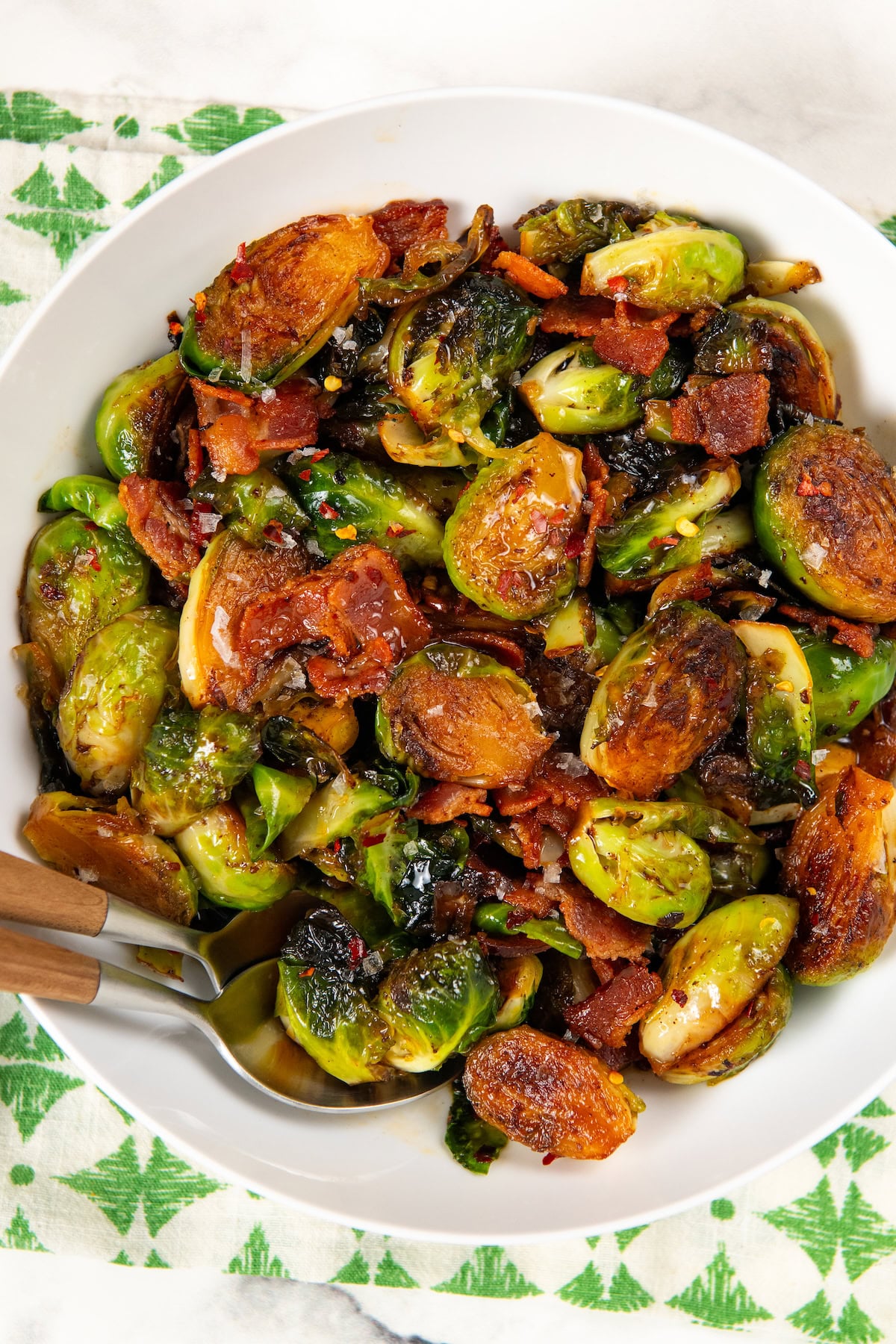 Hot Honey Brussels Sprouts with crispy bacon, caramelized sprouts, and a sweet-spicy honey drizzle. One skillet, ready in 25 minutes! A white bowl filled with hot honey Brussels sprouts mixed with crispy bacon pieces and topped with chili flakes and flaky sea salt.