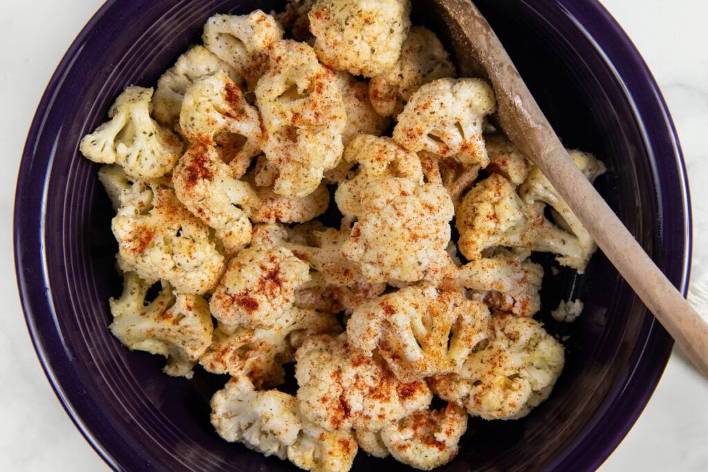 Parmesan Ranch Roasted Cauliflower recipe is a 5-ingredient side dish with crispy golden edges and big herby flavor. On the table in 30 minutes! A dark purple bowl filled with cauliflower florets sprinkled with ranch seasoning and paprika with a wooden spoon resting on the side of the bowl.