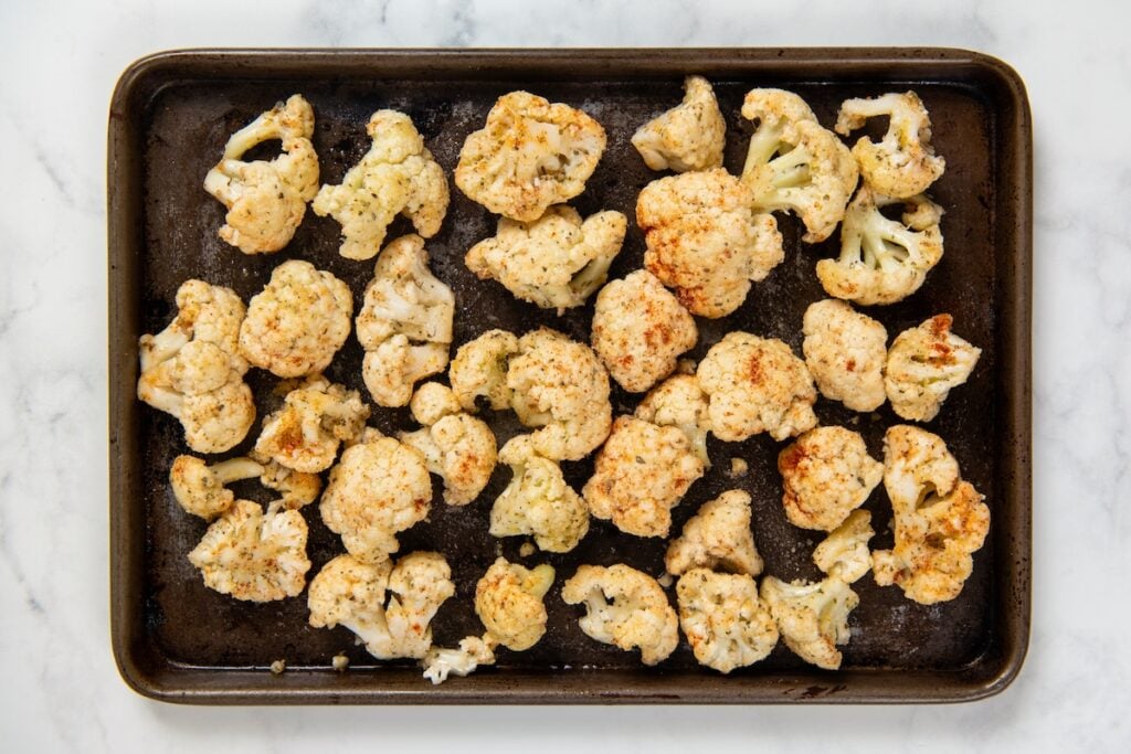 Parmesan Ranch Roasted Cauliflower recipe is a 5-ingredient side dish with crispy golden edges and big herby flavor. On the table in 30 minutes! Raw cauliflower florets seasoned with spices and spread evenly on a dark baking sheet.