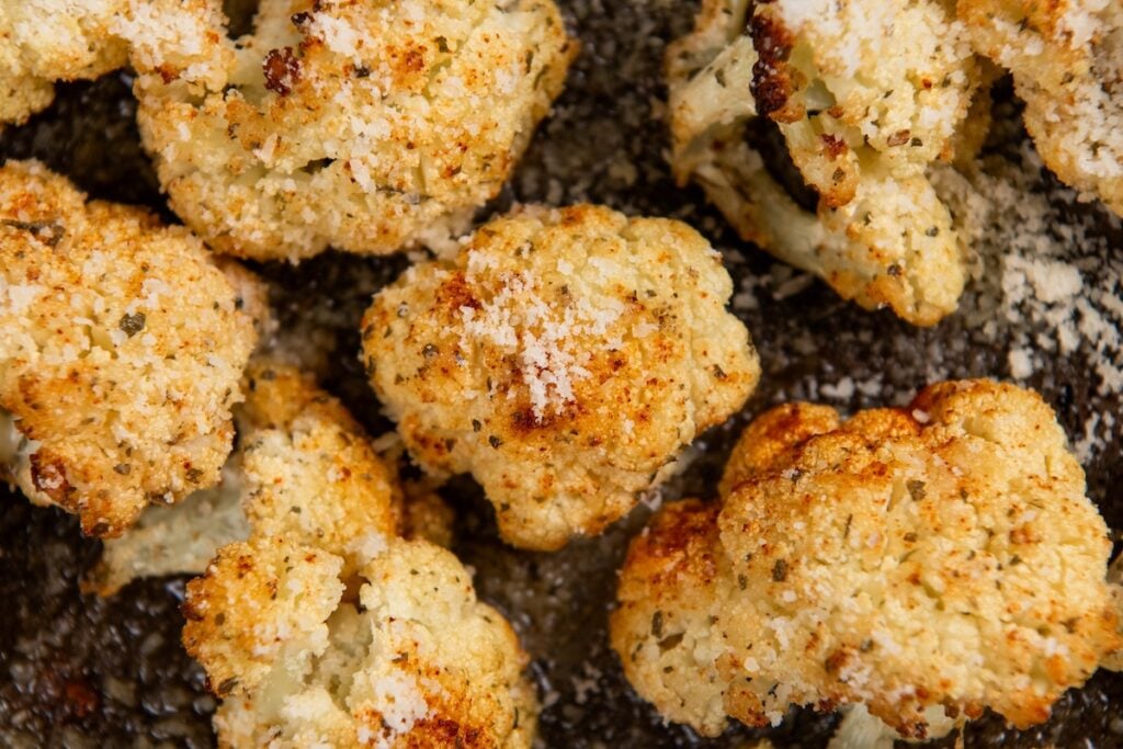 Parmesan Ranch Roasted Cauliflower recipe is a 5-ingredient side dish with crispy golden edges and big herby flavor. On the table in 30 minutes! Close-up of roasted Parmesan Ranch Roasted Cauliflower topped with grated cheese and spices, resting on a dark baking tray. The florets appear golden brown and crispy.