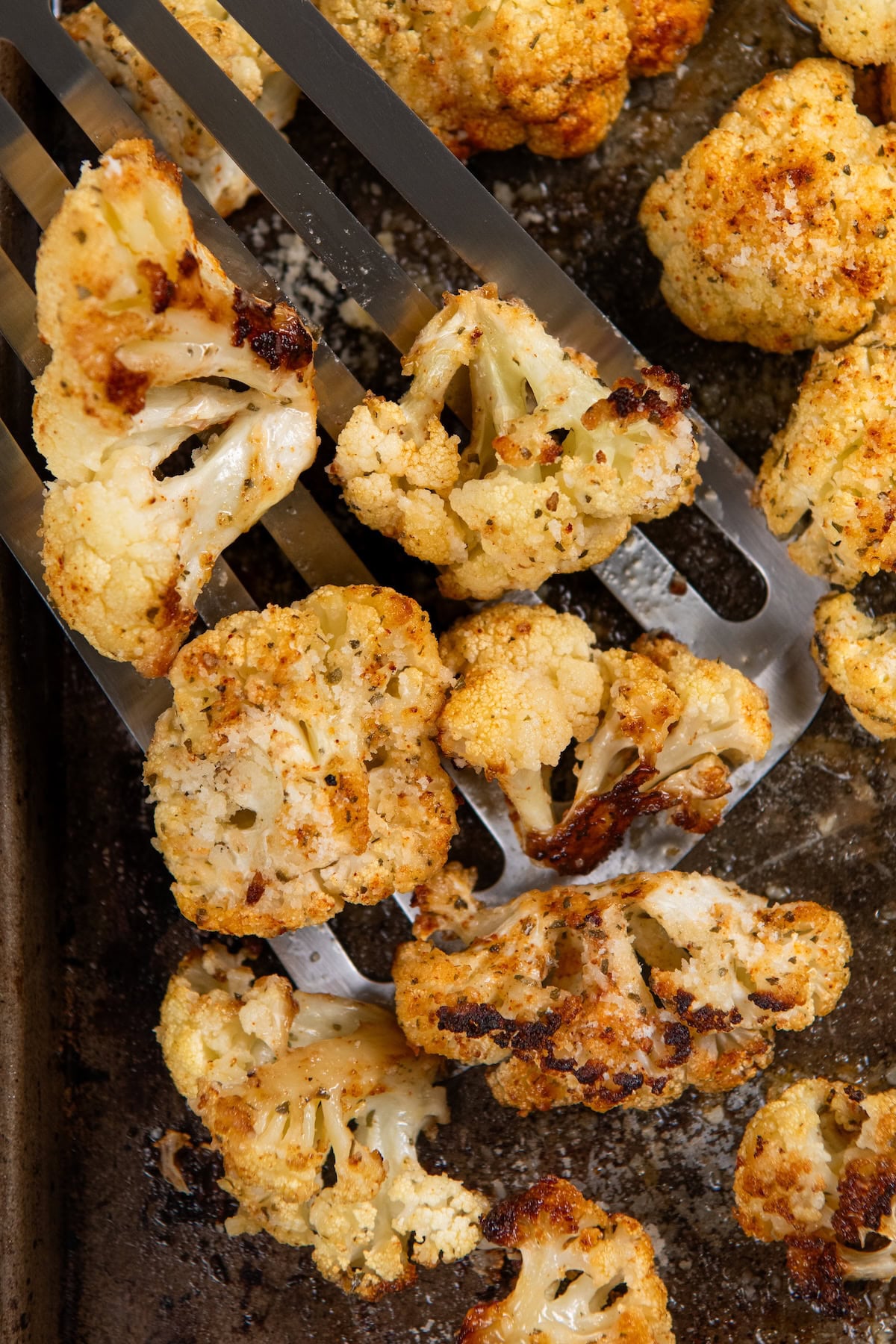 Parmesan Ranch Roasted Cauliflower recipe is a 5-ingredient side dish with crispy golden edges and big herby flavor. On the table in 30 minutes! Close-up of golden-brown roasted cauliflower florets on a baking sheet, with a metal spatula lifting some pieces. The cauliflower is seasoned, topped with parmesan cheese, and perfectly golden brown with crispy edges.