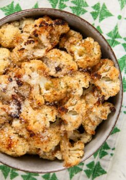 A bowl filled with parmesan ranch roasted cauliflower florets, sprinkled with grated cheese, sits on a white and green cloth.