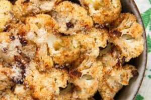 A bowl filled with parmesan ranch roasted cauliflower florets, sprinkled with grated cheese, sits on a white and green cloth.