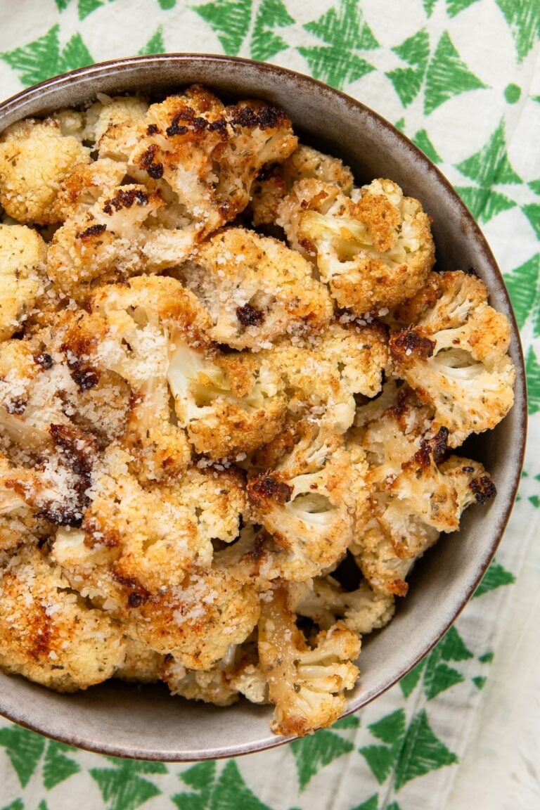 A bowl filled with parmesan ranch roasted cauliflower florets, sprinkled with grated cheese, sits on a white and green cloth.