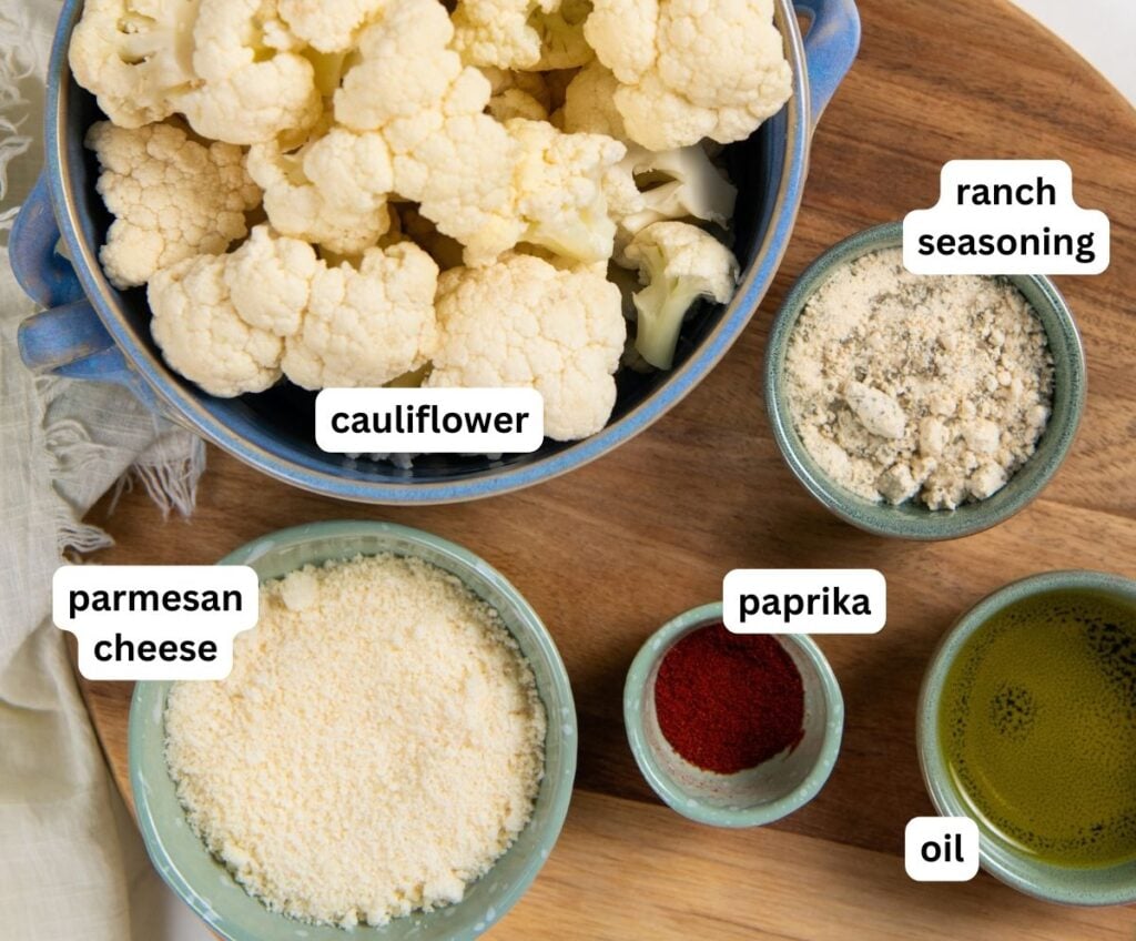 Parmesan Ranch Roasted Cauliflower recipe is a 5-ingredient side dish with crispy golden edges and big herby flavor. On the table in 30 minutes! A wooden board holds bowls of ingredients for a parmesan ranch roasted cauliflower recipe. Each bowl is labeled: cauliflower, ranch seasoning, parmesan cheese, paprika, and oil.
