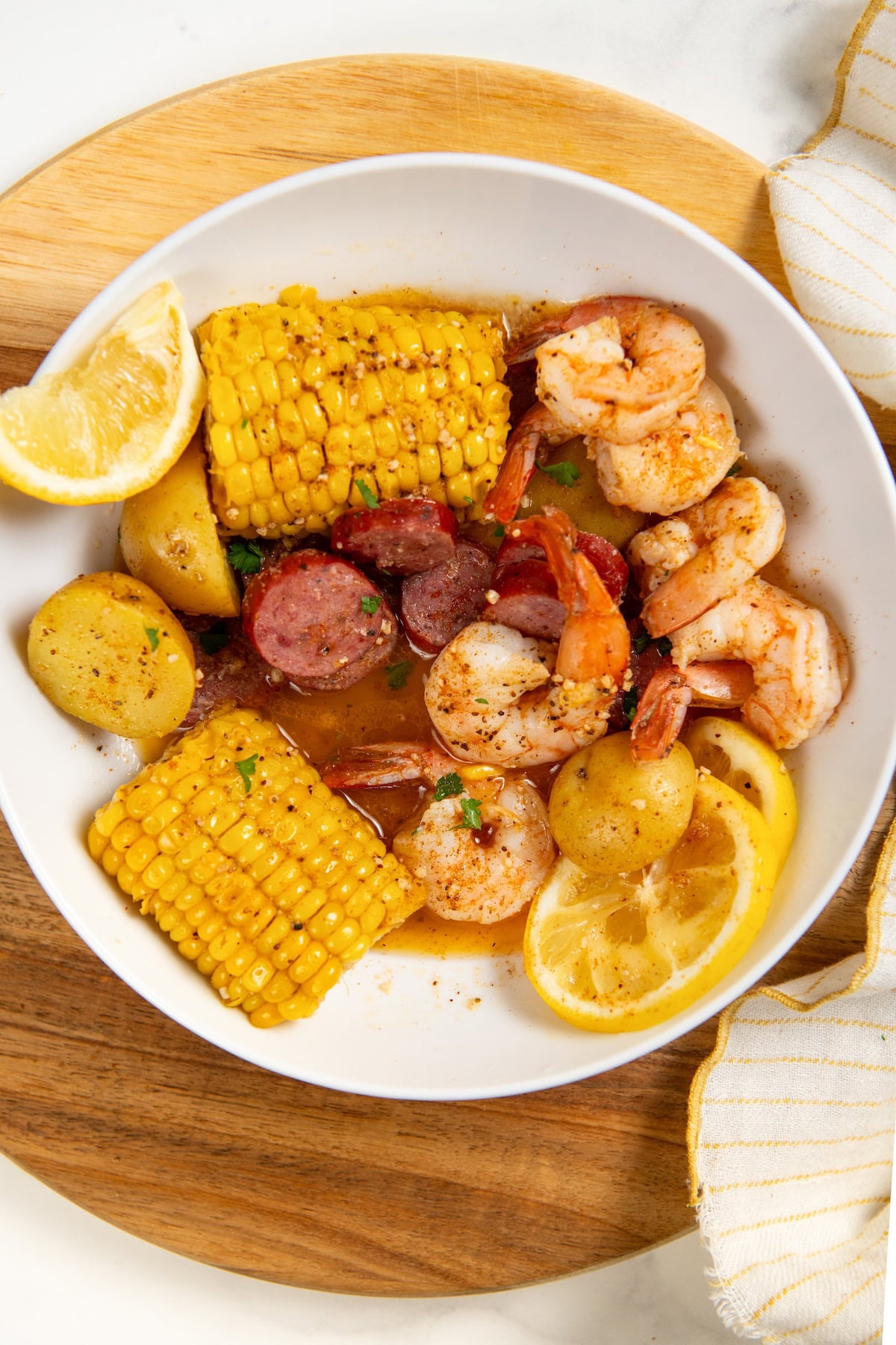 Shrimp Foil Packets bring all the flavor of a shrimp boil with garlic butter, sausage, corn, and potatoes. Ready in about 30 minutes! The content of the shrimp boil foil packets with sausage slices, corn on the cob, baby potatoes, and lemon wedges, garnished with herbs, served in a bowl on a wooden platter.