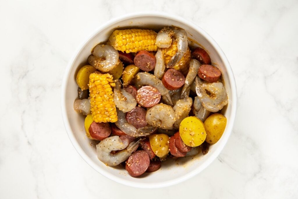 Shrimp Foil Packets bring all the flavor of a shrimp boil with garlic butter, sausage, corn, and potatoes. Ready in about 30 minutes! A white bowl filled with raw shrimp, sliced sausage, corn on the cob pieces, and small potatoes, all seasoned with spices, on a white marble surface.