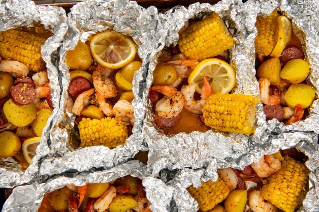 Shrimp Foil Packets bring all the flavor of a shrimp boil with garlic butter, sausage, corn, and potatoes. Ready in about 30 minutes! Shrimp foil packets filled with cajun shrimp, sliced sausage, corn on the cob, small potatoes, and lemon slices, all cooked together and arranged closely on a tray.