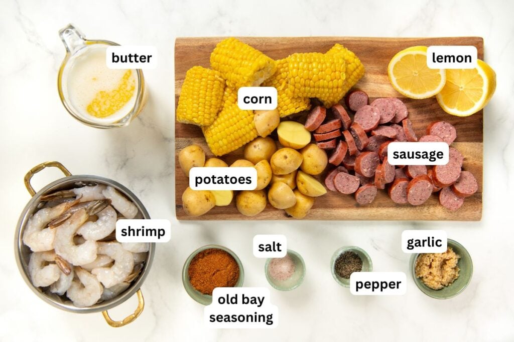 Labeled view of ingredients for shrimp foil packets: corn, potatoes, sausage, lemon, shrimp, butter, garlic, salt, pepper, and Old Bay seasoning, arranged on a white surface and wooden board.