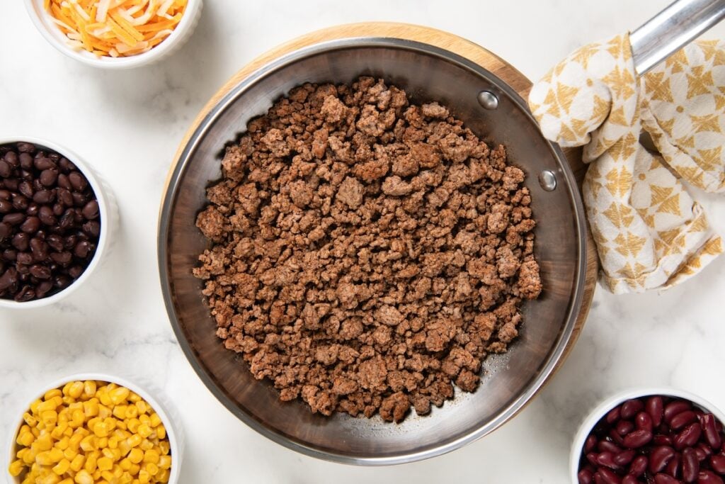 This hearty Ground Beef Taco Salad recipe combines all the best taco flavors in one bowl with a fresh cilantro lime dressing! Ready in 30 minutes. A skillet filled with cooked ground beef sits on a marble countertop, surrounded by small bowls of shredded cheese, black beans, kidney beans, and corn.