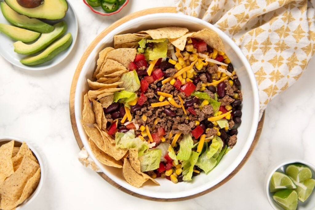 This hearty Ground Beef Taco Salad recipe combines all the best taco flavors in one bowl with a fresh cilantro lime dressing! Ready in 30 minutes. A bowl of ground beef taco salad with black beans, kidney beans, corn, lettuce, tomatoes, shredded cheese, and tortilla chips, surrounded by avocado slices, lime wedges, and extra chips on a white marble surface.