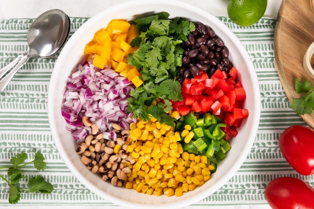 Best made in advance, Cowboy Caviar dip is fresh, colorful, and full of black beans, corn, and crunchy veggies in a tangy lime dressing. A white bowl filled with diced red onion, yellow bell pepper, black beans, red bell pepper, jalapeño, corn, black-eyed peas, and fresh cilantro sits on a striped cloth with spoons nearby.
