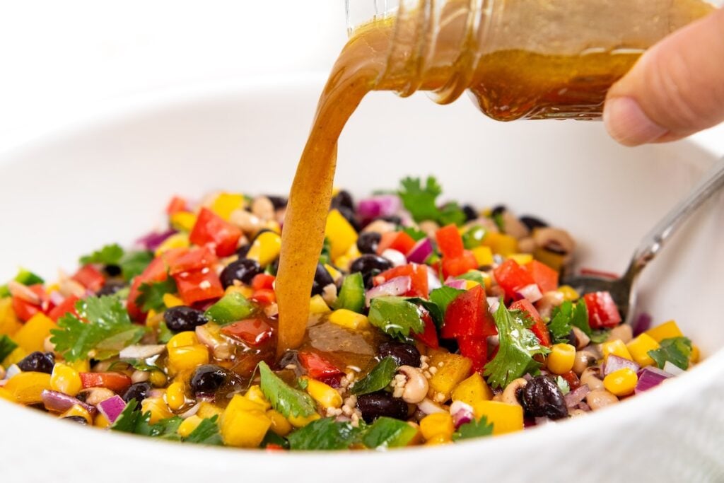 Best made in advance, Cowboy Caviar dip is fresh, colorful, and full of black beans, corn, and crunchy veggies in a tangy lime dressing. A hand pours cowboy caviar dressing from a jar onto a colorful mixture of black beans, corn, tomatoes, bell pepper, jalapeño, onions, and cilantro in a white bowl.