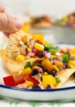 A hand holds a tortilla chip topped with colorful cowboy caviar over a white plate with more chips and dip.
