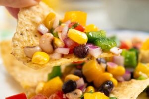 A hand holds a tortilla chip topped with colorful cowboy caviar over a white plate with more chips and dip.