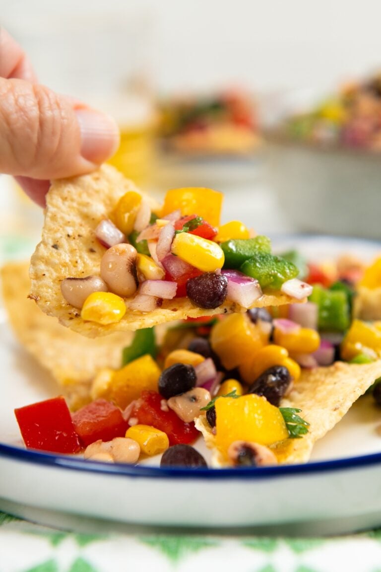 A hand holds a tortilla chip topped with colorful cowboy caviar over a white plate with more chips and dip.