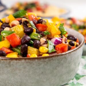 A bowl of colorful cowboy caviar dip with black beans, corn, red and yellow bell peppers, red onion, and chopped parsley, sitting on a patterned cloth.