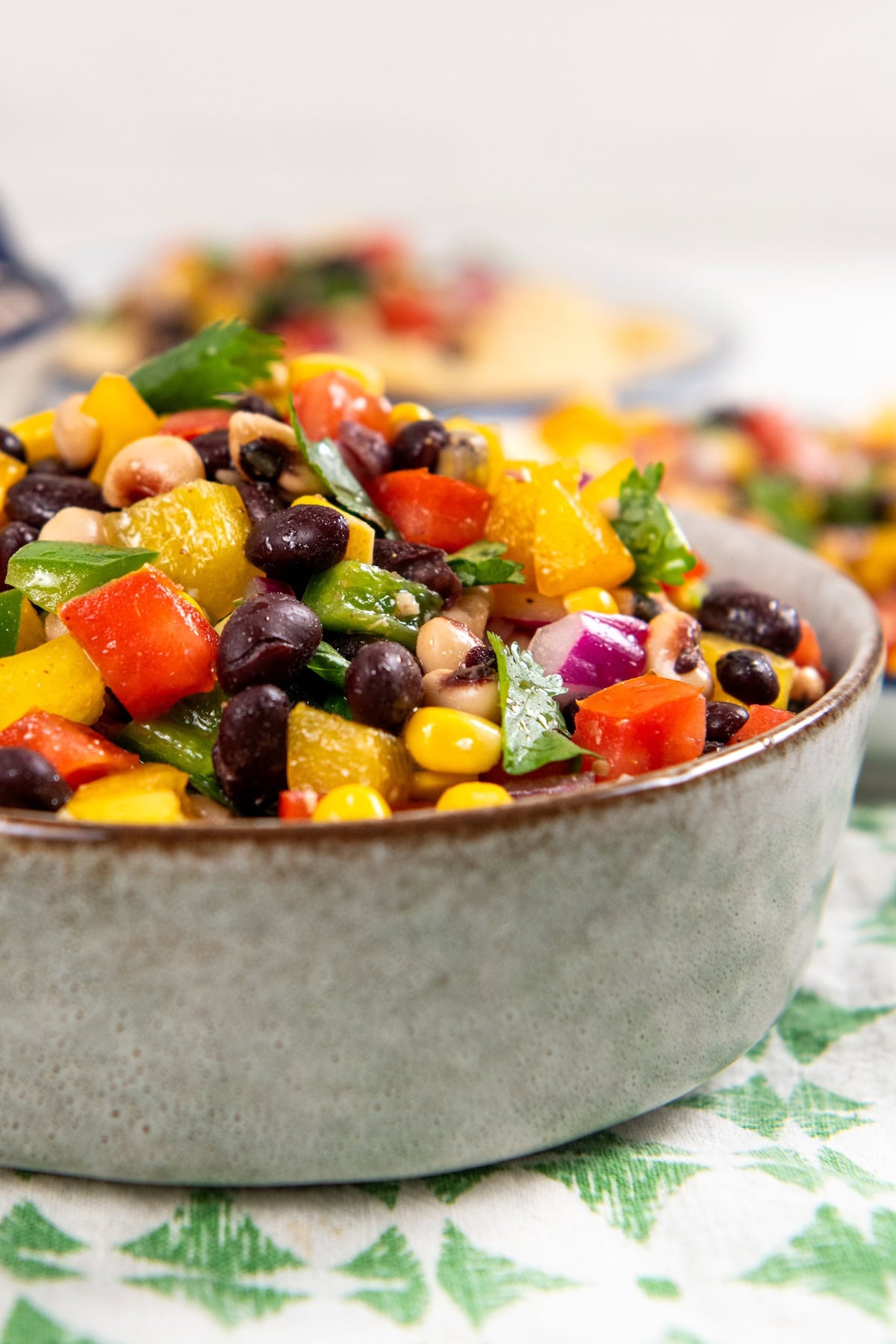 Best made in advance, Cowboy Caviar dip is fresh, colorful, and full of black beans, corn, and crunchy veggies in a tangy lime dressing. A bowl of colorful cowboy caviar dip with black beans, corn, red and yellow bell peppers, red onion, and chopped parsley, sitting on a patterned cloth.