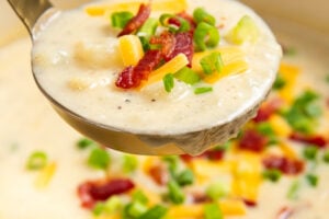 A ladle lifts creamy potato bacon soup topped with shredded cheese, crispy bacon bits, and chopped green onions from a blue pot.