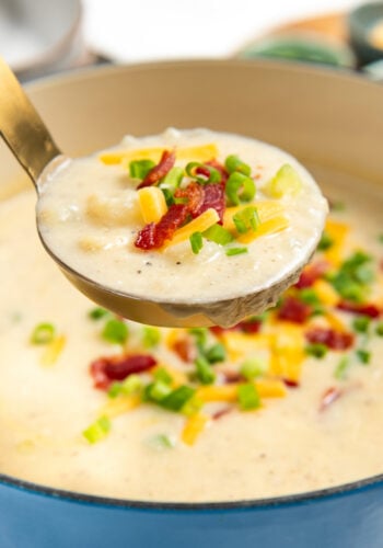A ladle lifts creamy potato bacon soup topped with shredded cheese, crispy bacon bits, and chopped green onions from a blue pot.