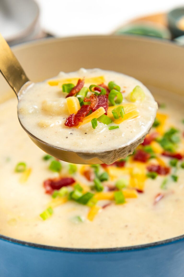 A ladle lifts creamy potato bacon soup topped with shredded cheese, crispy bacon bits, and chopped green onions from a blue pot.