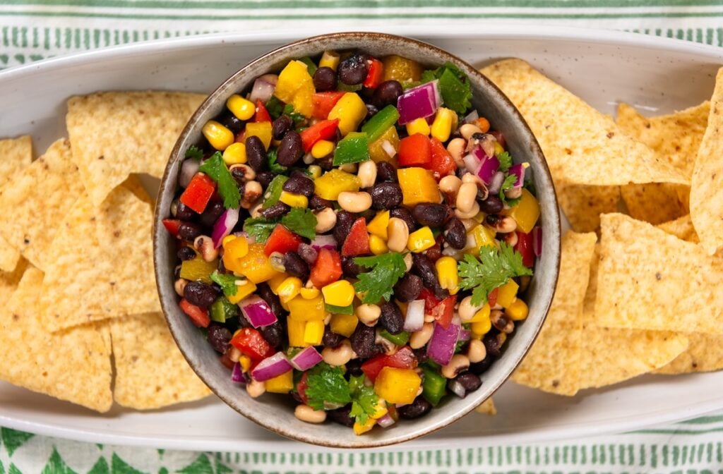 Best made in advance, Cowboy Caviar dip is fresh, colorful, and full of black beans, corn, and crunchy veggies in a tangy lime dressing. A bowl of colorful cowboy caviar dip with diced peppers, onions, and cilantro, surrounded by tortilla chips on a rectangular white platter.