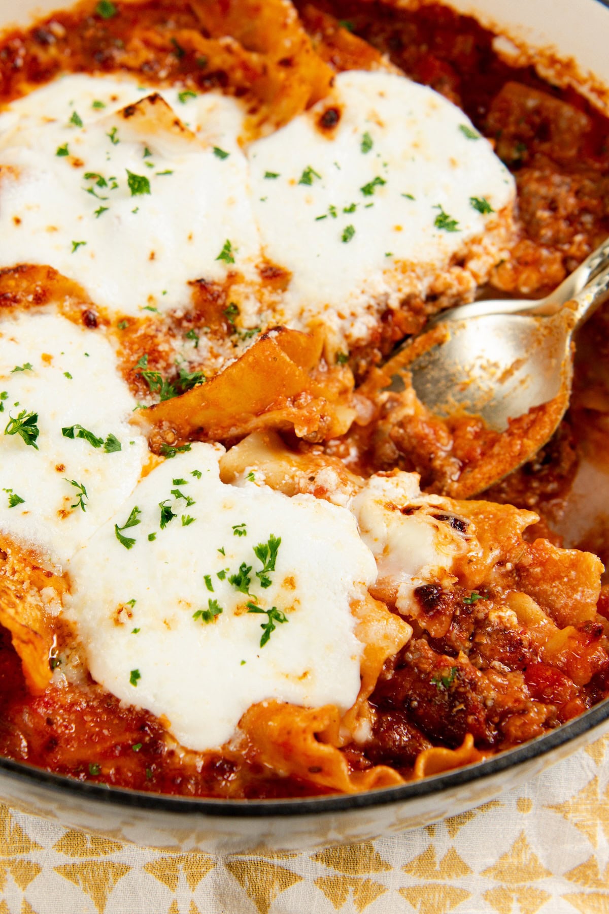 Skillet Lasagna is a quick one pan spin on the classic. No boiling, no layering, and a golden broiled cheese top in about 30 minutes! A close-up of baked skillet lasagna, topped with melted cheese and parsley, with a serving spoon lifting a portion.