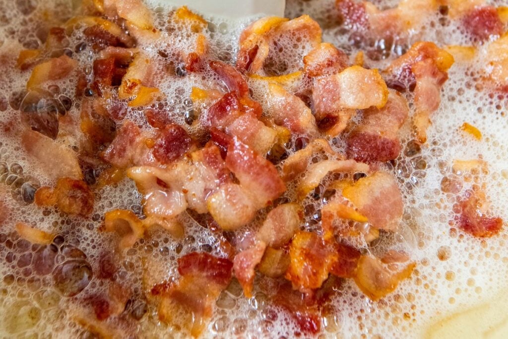 Crispy bacon pieces sizzling in hot grease, surrounded by bubbling foam as they cook in a pan.