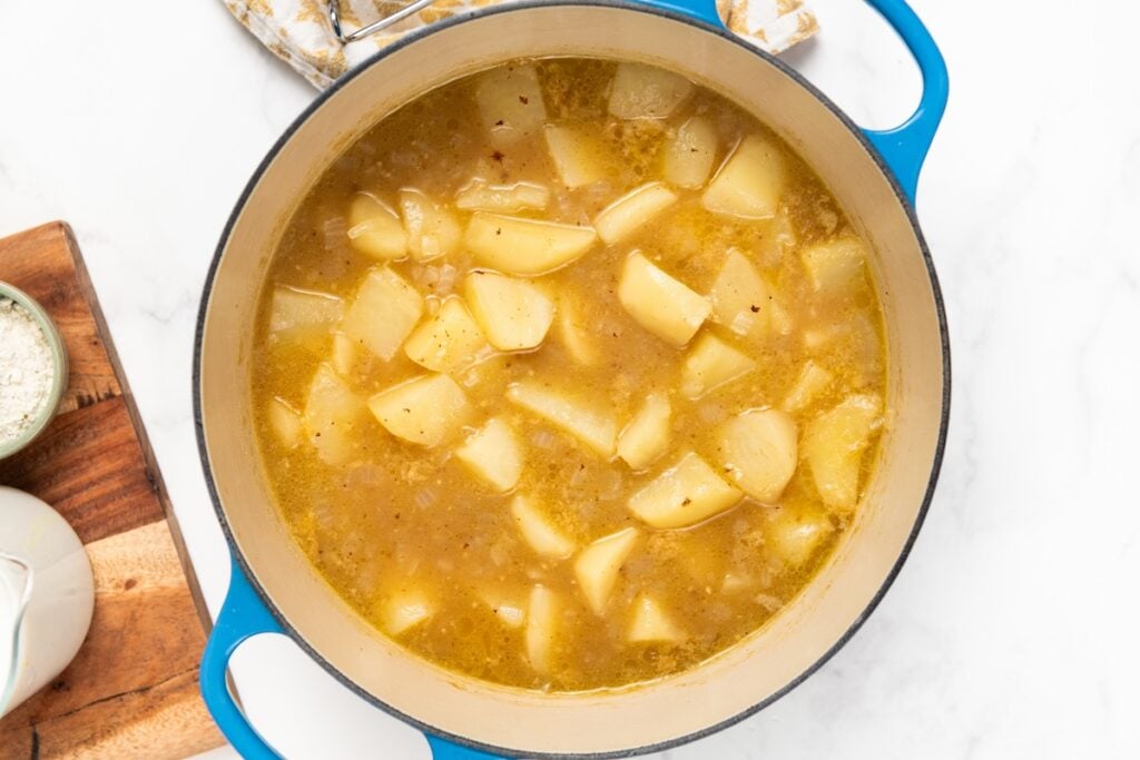 A blue pot filled with cooked potatoes in broth sits on a white surface, with a cutting board and a small bowl of salt nearby.