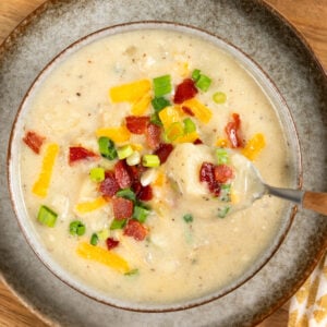 A bowl of creamy bacon potato soup topped with shredded cheese, chopped bacon, and green onions sits on a wooden board. A spoon is lifting some soup from the bowl.