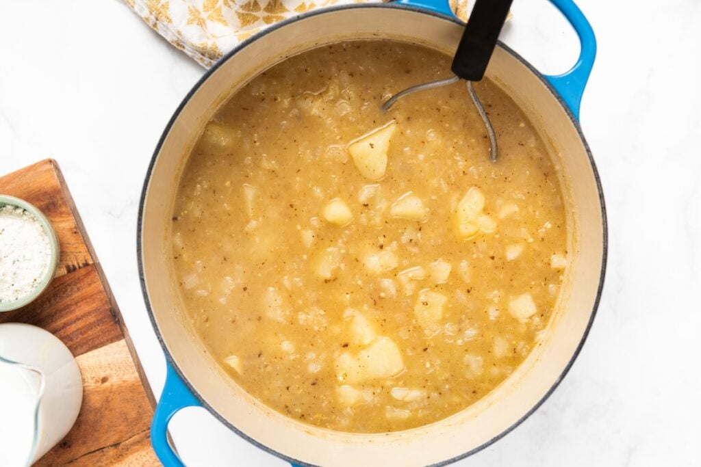 A blue pot filled with chunky potato soup sits on a white surface. A potato masher rests inside the pot.