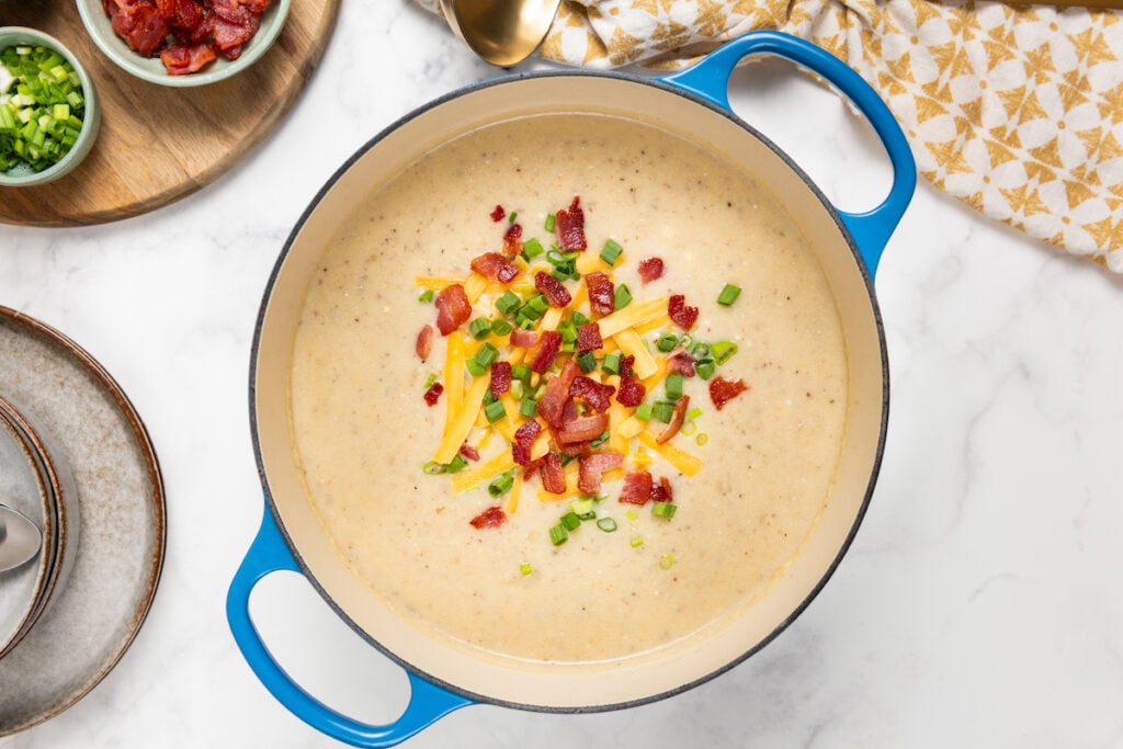A pot of creamy potato and bacon soup topped with shredded cheese, bacon bits, and chopped green onions, placed on a marble counter next to a small bowl of toppings and a gold spoon.