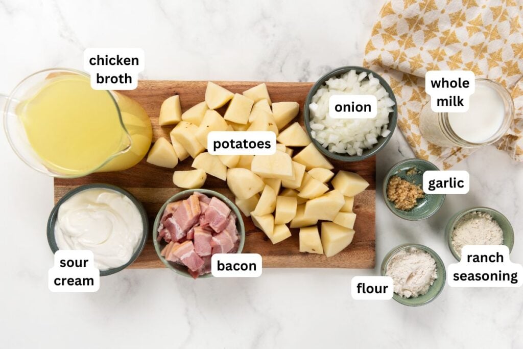 Thick, smoky, and creamy, Bacon Potato Soup is made with bacon drippings and topped with crispy bacon. One pot and ready in under an hour! Ingredients for potato and bacon soup recipe arranged on a cutting board and labeled: chopped potatoes, bacon, onion, garlic, ranch seasoning, flour, sour cream, chicken broth, and whole milk.