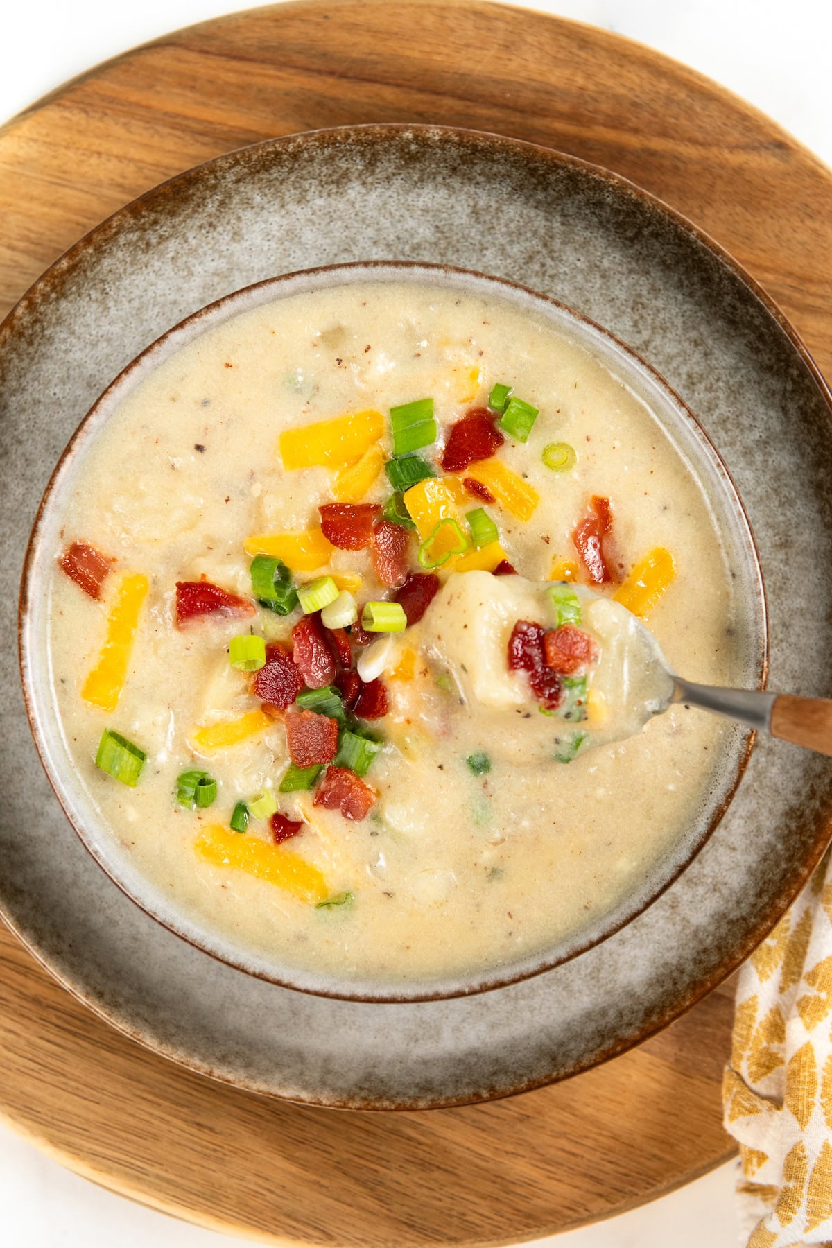 Thick, smoky, and creamy, Bacon Potato Soup is made with bacon drippings and topped with crispy bacon. One pot and ready in under an hour! A bowl of creamy bacon potato soup topped with shredded cheese, chopped bacon, and green onions sits on a wooden board. A spoon is lifting some soup from the bowl.