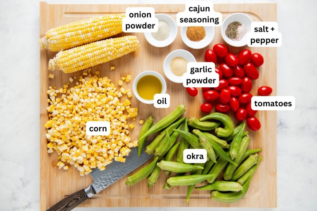 Roasted Okra and Tomatoes with Corn is a crispy spin on the Southern classic. One sheet pan, 10 minutes of prep, and zero slimy okra! Chopped and whole corn, okra, tomatoes, small bowls of oil, onion powder, garlic powder, Cajun seasoning, salt, and pepper are on a wooden cutting board with a knife.