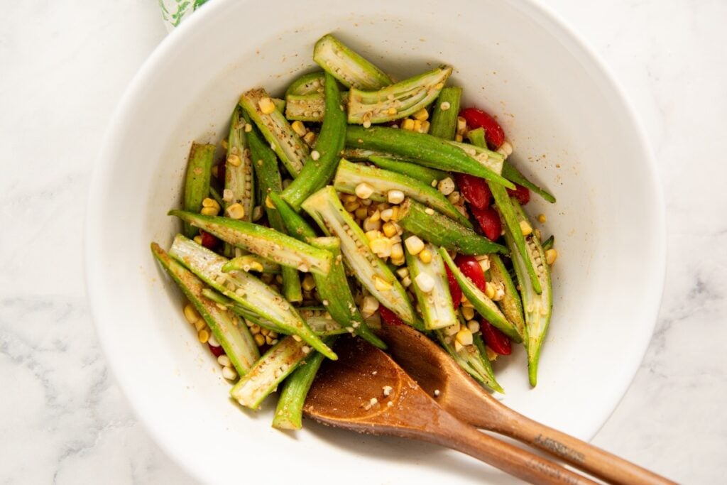 Roasted Okra and Tomatoes with Corn is a crispy spin on the Southern classic. One sheet pan, 10 minutes of prep, and zero slimy okra! A white bowl contains sliced okra, corn kernels, and cherry tomatoes, mixed with spices. Two wooden utensils rest inside the bowl on a marble surface.