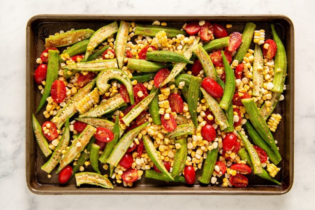 Roasted Okra and Tomatoes with Corn is a crispy spin on the Southern classic. One sheet pan, 10 minutes of prep, and zero slimy okra! A baking tray with sliced okra, cherry tomatoes, and corn kernels, all seasoned and ready to be roasted, on a white marble surface.