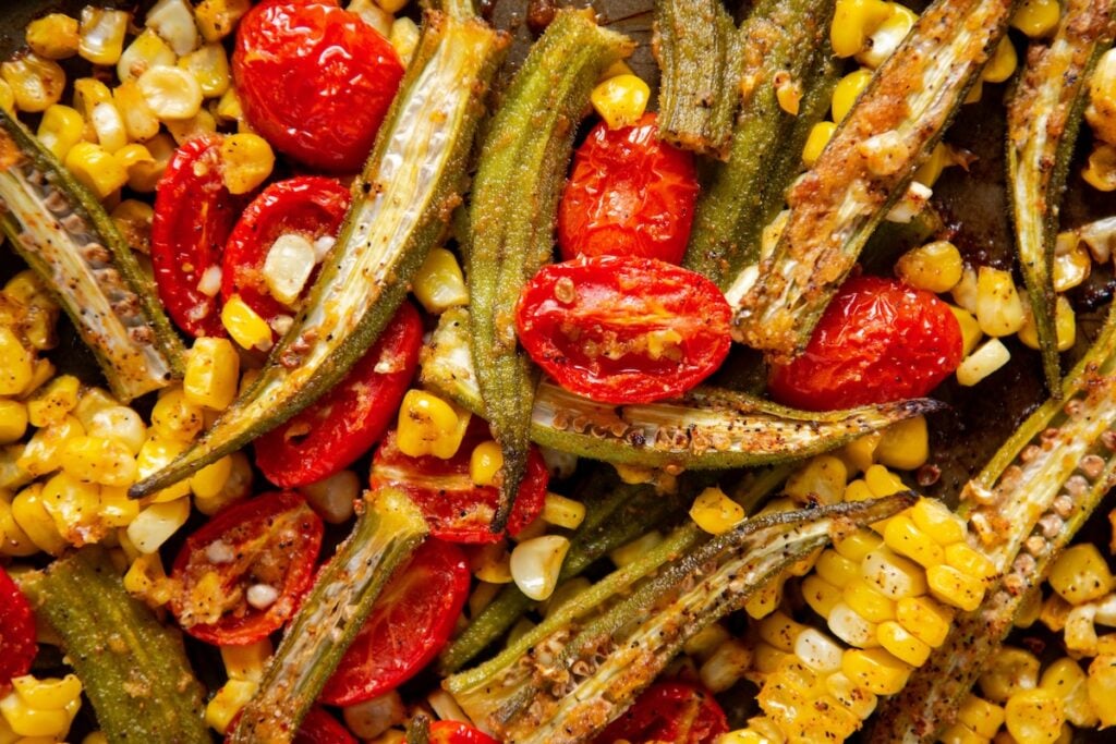 Roasted Okra and Tomatoes with Corn is a crispy spin on the Southern classic. One sheet pan, 10 minutes of prep, and zero slimy okra! Close-up of roasted okra and tomatoes with corn that has been seasoned with spices. The vegetables are slightly charred, vibrant, and arranged together, showing texture and color variety in a cooked vegetable medley.