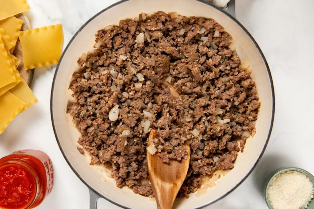 Skillet Lasagna is a quick one pan spin on the classic. No boiling, no layering, and a golden broiled cheese top in about 30 minutes! A pot filled with cooked ground sausage and chopped onions, stirred with a wooden spoon. Uncooked lasagna noodles, a jar of tomato sauce, and a small bowl of grated cheese are nearby.
