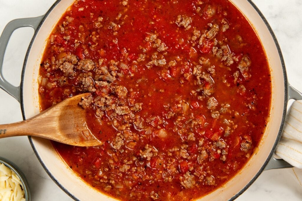 Skillet Lasagna is a quick one pan spin on the classic. No boiling, no layering, and a golden broiled cheese top in about 30 minutes! A pot of hearty meat sauce with ground meat, onion, garlic, and marinara sauce is being stirred with a wooden spoon.
