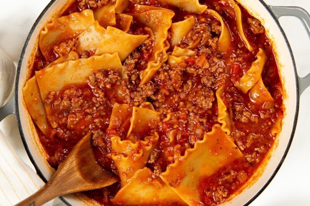 Skillet Lasagna is a quick one pan spin on the classic. No boiling, no layering, and a golden broiled cheese top in about 30 minutes! A pot of lasagna noodles cooking with ground meat and marinara sauce is being stirred with a wooden spoon.