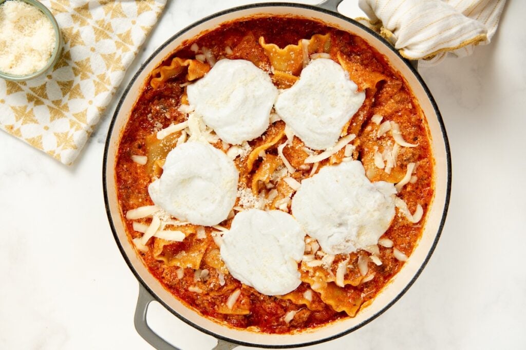 Skillet Lasagna is a quick one pan spin on the classic. No boiling, no layering, and a golden broiled cheese top in about 30 minutes! A round skillet of lasagna with wavy pasta, tomato sauce, shredded cheese, and six dollops of cottage cheese, sitting on a white surface next to a bowl of grated cheese and a cloth napkin.