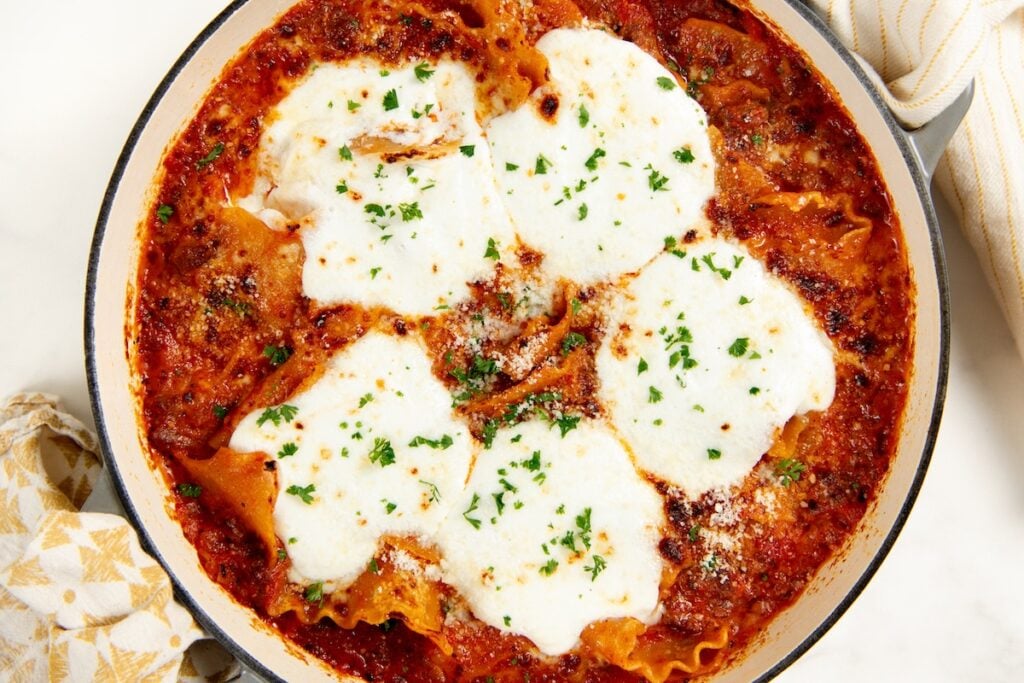 Skillet Lasagna is a quick one pan spin on the classic. No boiling, no layering, and a golden broiled cheese top in about 30 minutes! A baked lasagna skillet with layers of red tomato sauce, melted cheese, and sprinkled parsley on top, placed next to a patterned kitchen towel.