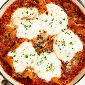 A baked lasagna skillet with layers of red tomato sauce, melted cheese, and sprinkled parsley on top, placed next to a patterned kitchen towel.
