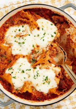 A close-up of a skillet lasagna topped with melted cheese and sprinkled with herbs, with a serving spoon partially inside the dish.