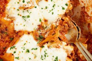 A close-up of a skillet lasagna topped with melted cheese and sprinkled with herbs, with a serving spoon partially inside the dish.