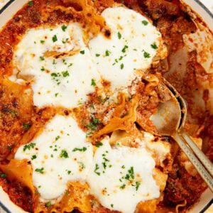 A close-up of a skillet lasagna topped with melted cheese and sprinkled with herbs, with a serving spoon partially inside the dish.